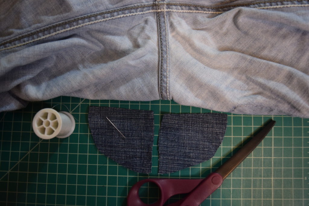 the back crotch of jeans shown with a spool of thread, a needle, scissors, and two dark triangular denim patche