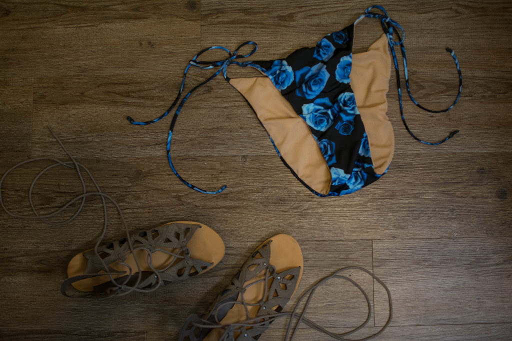 a flat lay of blue rose print string bikini bottoms with side ties on a hardwood floor with strappy sandals