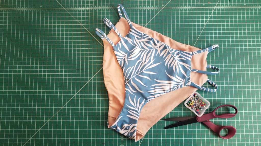 a pair of light blue bikini bottoms with three straps on each side laying on a cutting mat with a pair of scissors and container of pins